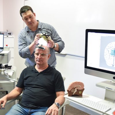A man sits on a chair as a second man stands behind him using an ultrasound device on his head.